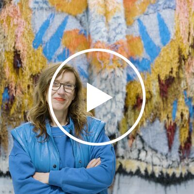 Artist Caroline Achaintre, a middle-aged woman with long wavy brown hair, wearing glasses and a powder blue sweatshirt and sleeveless jacket, stands in front of one of her multi-coloured woven artworks