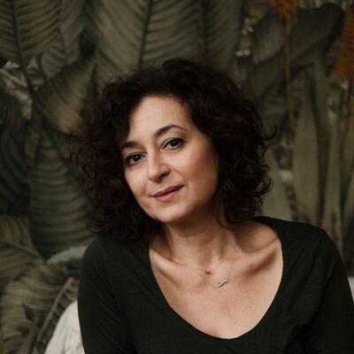 Author Ece Temelkuran sits leaning against a sofa with a green background in a black top