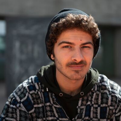 Portrait of a young man with curly hair and a mustache wearing a black beanie, a plaid overshirt, and a dark hoodie, looking directly at the camera.