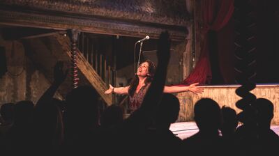A female singer with long dark hair performs on a dimly lit, vintage stage with her arms wide open, singing passionately to a silhouetted audience.