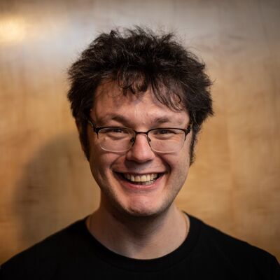 Close-up of a smiling man with glasses and messy dark hair, posing against a light-brown wooden paneled wall.