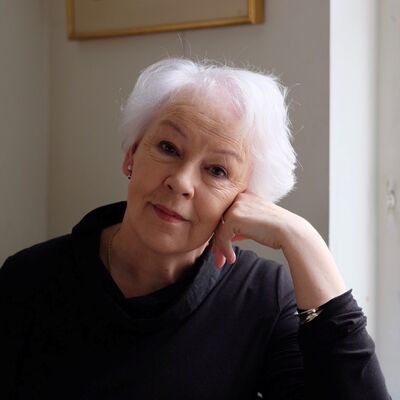 A woman with short white hair rests her head on her hand in front of a framed landscape painting on a white wall.