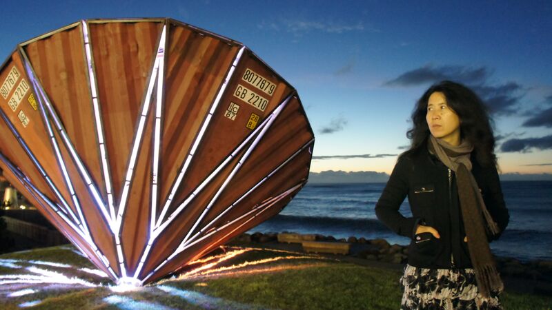 The artist Yin Xiuzhen stands with her hands in her jacket pockets in front of an outdoor sculpture piece