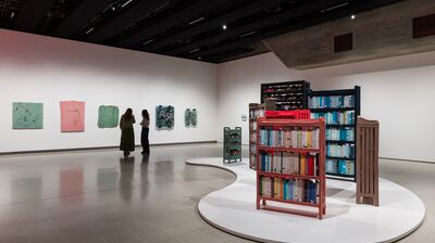 Two people admire artwork on a gallery wall. In the foreground, bookshelves with books made of cloth on them