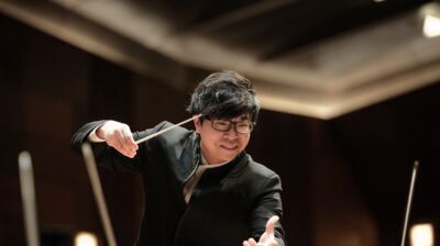 Kahchun Wong conducting with his baton, using both arms and smiling looking down toward the orchestra