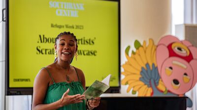 Charlotte, a Black women artist smiles as she reads her own poetry in front of a yellow screen.