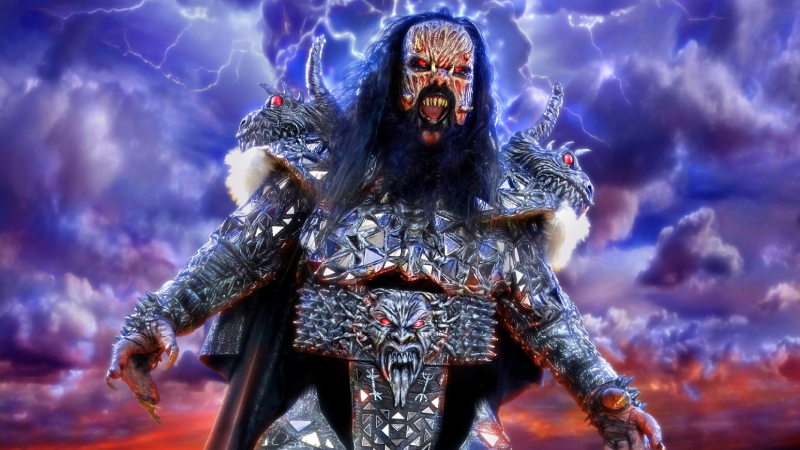 Mr. Lordi dressed as a grotesque zombie wearing shiny metallic armour, stood in front of an apocalyptic landscape