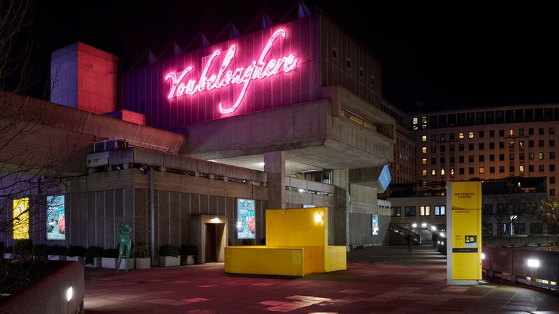 A brutalist building at night, featuring a large pink neon sign that reads "You belong here" above a yellow structure.