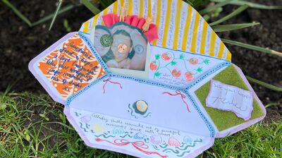 Handmade paper craft with drawings and photos sits on grass in front of several bright yellow daffodils.