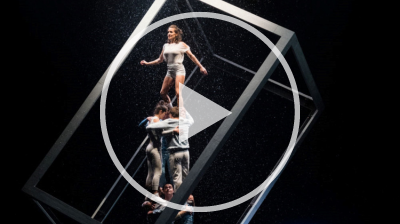 A dramatic acrobatic performance on a dark stage with simulated rain. A woman stands atop a human pyramid inside a large, tilted white cube frame, while a man plays a piano.