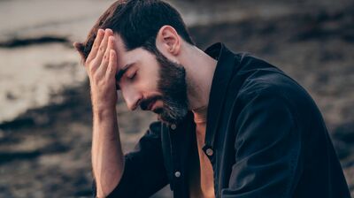 A man wearing a blue shirt sitting on a sunset beach with his palm against his forehead