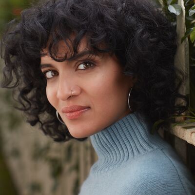 Close-up of a woman with curly dark hair and a nose ring, wearing a blue turtleneck in front of green foliage.