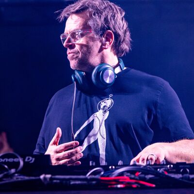 A DJ with glasses and headphones around his neck performs behind Pioneer DJ decks under atmospheric purple stage lighting.