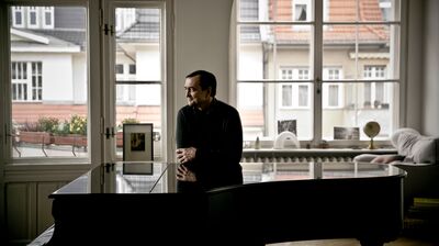 A man standing in a room with big windows leaning on a grand piano