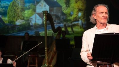 A white man wearing a white shirt, reading from a lectern.