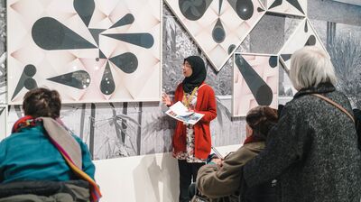 A guide in a red cardigan and hijab speaks to visitors in front of abstract geometric artwork.