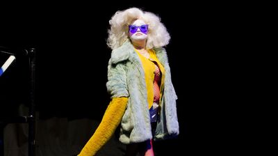 A performer in a large blonde wig, wearing a yellow top and a white fur coat on a dark stage