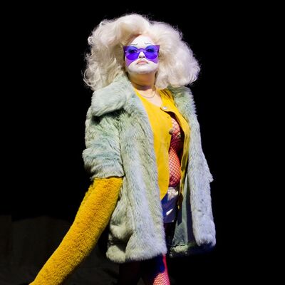 A performer in a large blonde wig, wearing a yellow top and a white fur coat on a dark stage