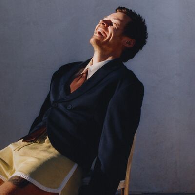 Harry Styles leans back in a chair, wearing a navy blue suit jacket, white shirt, brown silk tie and yellow shorts.