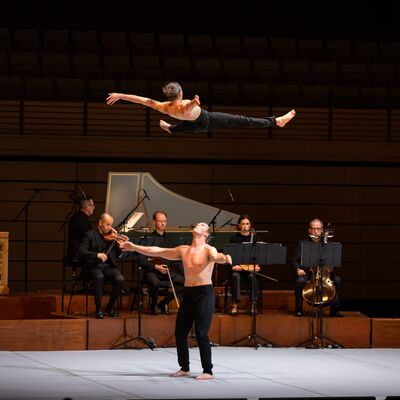 One shirtless acrobat thrown in the air above another acrobat who stands ready to catch them. Behind them are five musicians sat in front of a harpsichord and harmonium.