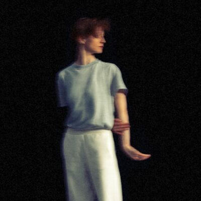A blurry, grain-textured image of a woman with red hair in a blue shirt and white pants, posing against a black background.