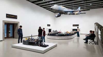 People view an art installation featuring a soft-sculpture airplane hanging above luggage carousels with miniature cities.