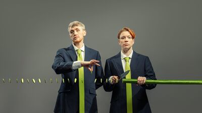 Two performers wearing blue suits and floor length green ties, cutting a very long cucumber with slices floating in the air