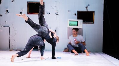 Two performers dressed in black entangled together, with a performer watching on with a microwave balanced on his head