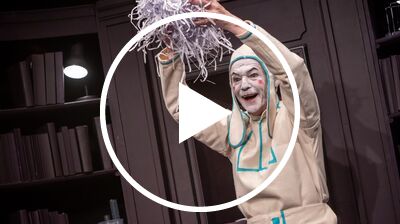 A performer in white face paint and a cream-colored hood joyfully tosses shredded paper into the air on a library stage set.