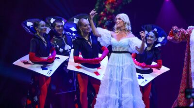 A performer in a ruffled light blue gown stands amidst dancers costumed as giant playing cards on a purple-lit stage.