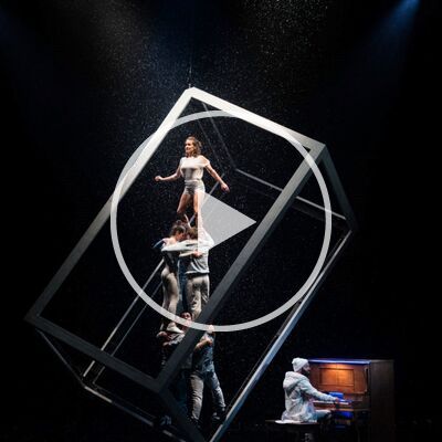 A dramatic acrobatic performance on a dark stage with simulated rain. A woman stands atop a human pyramid inside a large, tilted white cube frame, while a man plays a piano