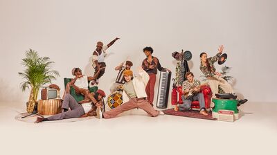 Group of performers posing with instruments, records and houseplants on a white backdrop