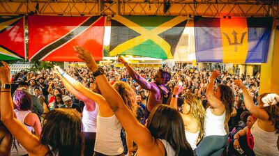Dancers with raised arms lead a large crowd under Caribbean flags, including Guyana, Trinidad, Jamaica, and Barbados.