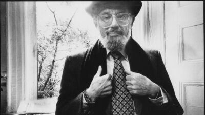 Black and white portrait of a bearded man with glasses and a hat, adjusting his patterned tie while looking at the camera.