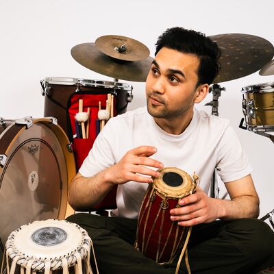A man in a white t-shirt sits on the floor surrounded by a drum kit and several traditional hand drums.