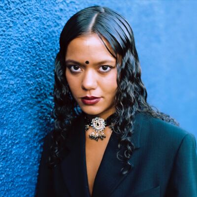 A woman with long dark hair in a black blazer leans on a bright blue wall