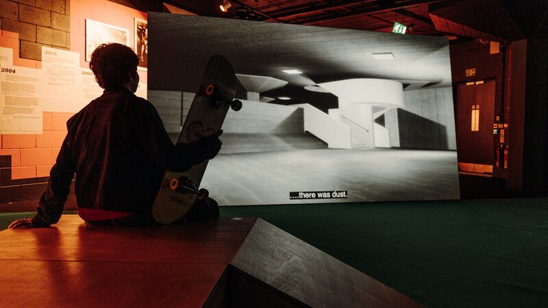 A silhouette of a person holding a skateboard sits on a ramp, watching a large film projection of concrete architecture.