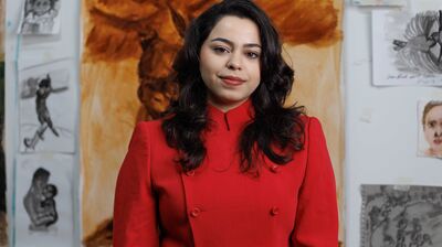 A woman with long wavy dark hair wears a double-breasted red blazer, standing in front of an art studio wall with sketches.