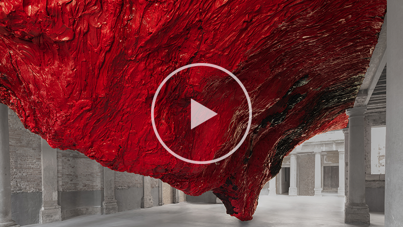 Large red textured sculpture hanging down from the ceiling to the floor in a space with stone floors and pillars