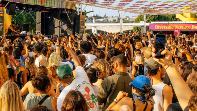 A dense crowd dances at an outdoor Riverside Terrace Bar under a canopy of colorful hexagonal decorations.