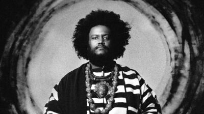 Black and white portrait of a man with an afro in a striped robe and ornate necklace, standing before a circular backdrop.