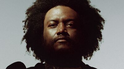 Low-angle portrait of a man with a large afro and beard wearing an ornate black garment with gold floral patterns.