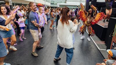 A crowd dances at an outdoor festival as a performer in a bright feathered costume dances on a stage in the background.