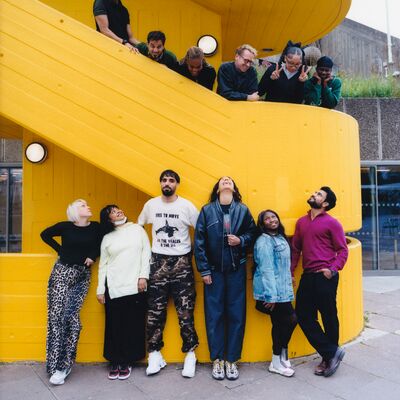 A diverse group of twelve people pose playfully on and underneath a bright yellow external concrete staircase outdoors