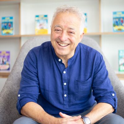 Frank Cottrell Boyce wearing a blue shirt sitting in front of a shelf of books.