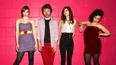 Four people photographed in front of a hot pink brick wall. They wear indie sleaze clothes.