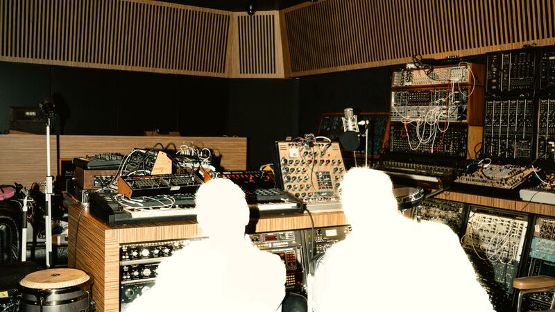 Two bright, overexposed figures sit in a recording studio filled with complex synthesizers and audio equipment.