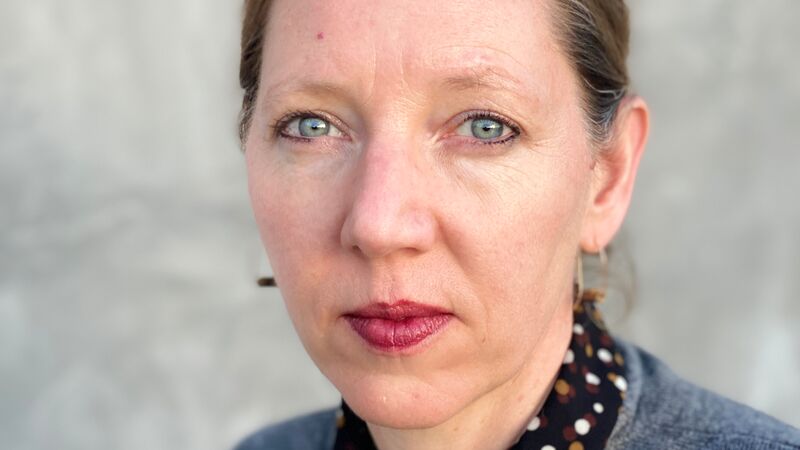 A woman with blue eyes and light hair looks directly at the camera, wearing a grey sweater over a polka-dot collared shirt.