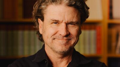 Dave Eggers wearing a black shirt standing in front of a bookshelf.