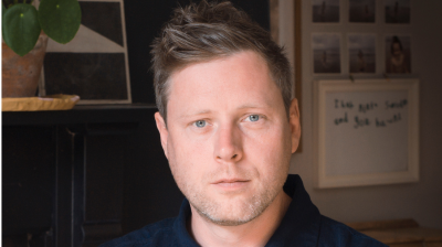 Max porter wears a blue workman's jacket buttoned up to the collar and looks straight at camera. In the background are polaroid images and sketchings.